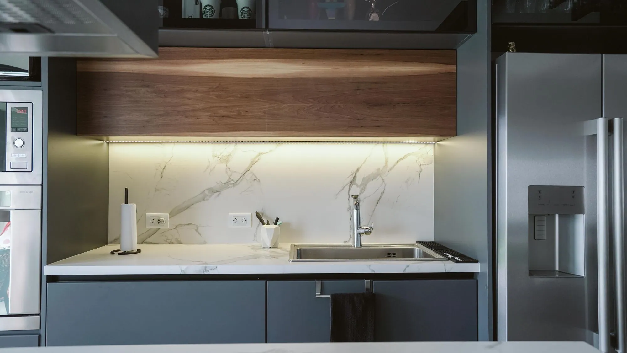 Graphite slab fronts under a thin white counter, with an LED strip washing a book-matched marble splash
