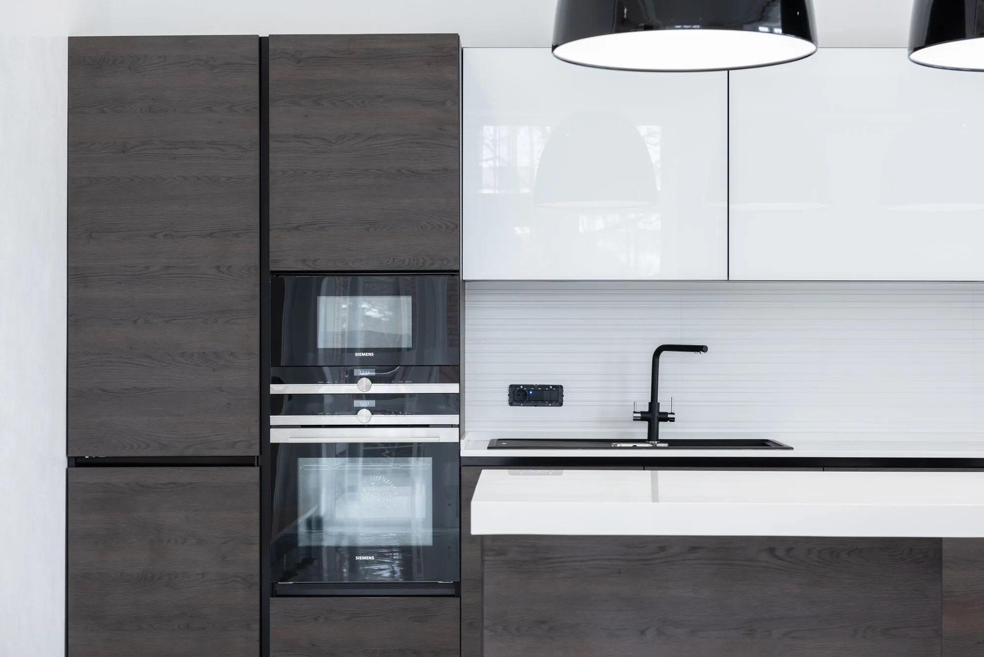 Dark oak handleless fronts beside white gloss uppers, with a tall bank carrying two built-in ovens