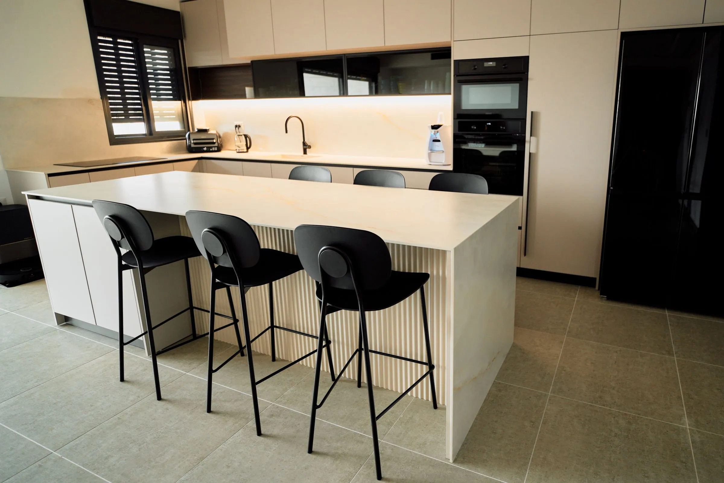 A long island with a fluted front and a pale stone top, facing handleless tall units and built-in ovens