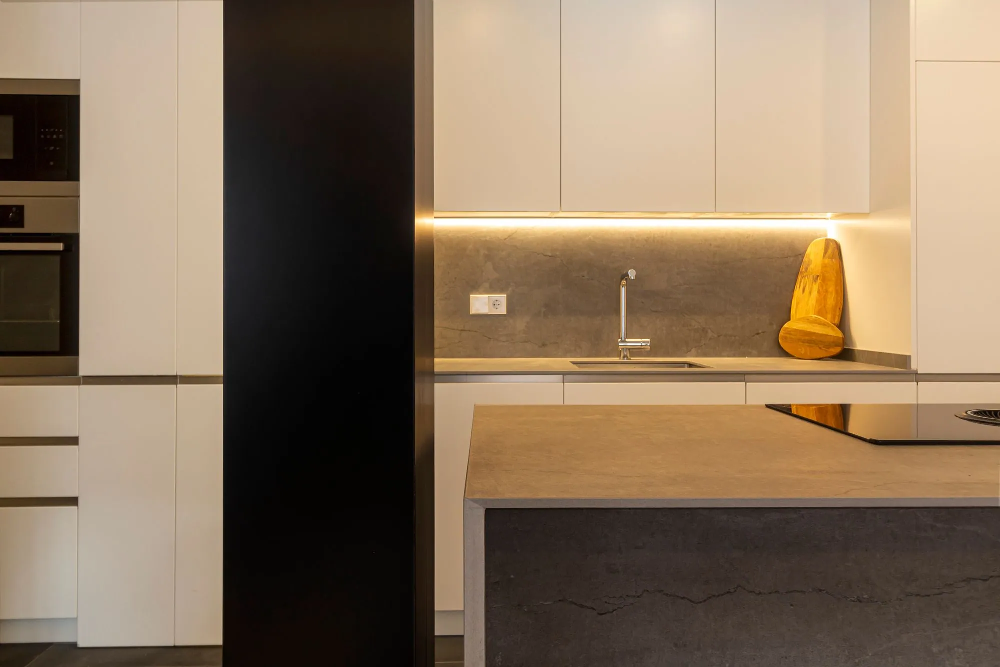 Super-matte white tall units and wall cabinets above a stone island, with a lit channel pull running under the upper cabinets