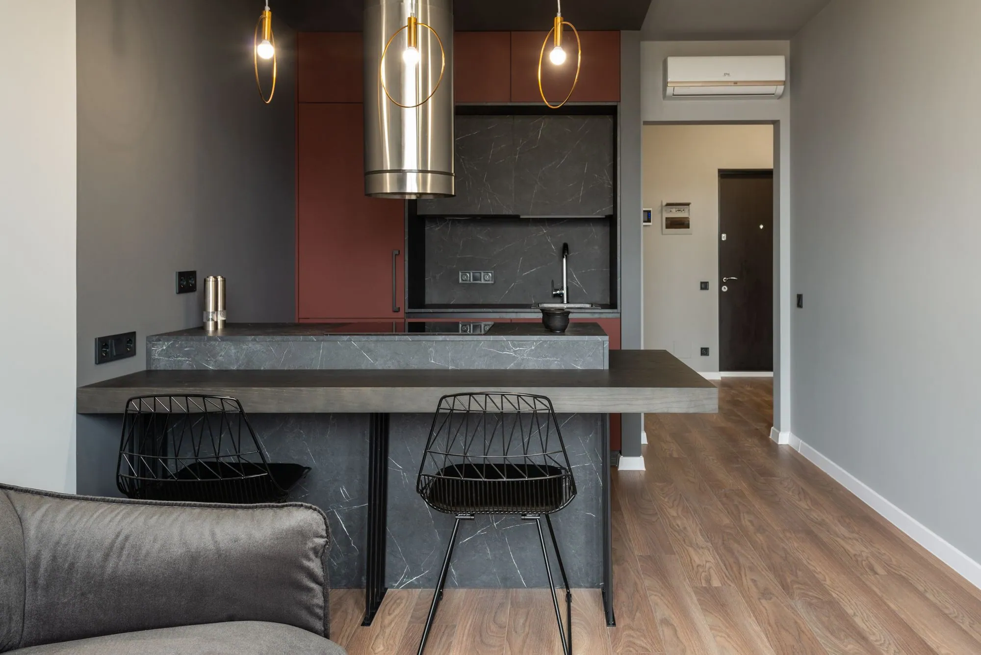 Compact bar peninsula in stone with a raised timber counter and two stools, against a matte tall bank
