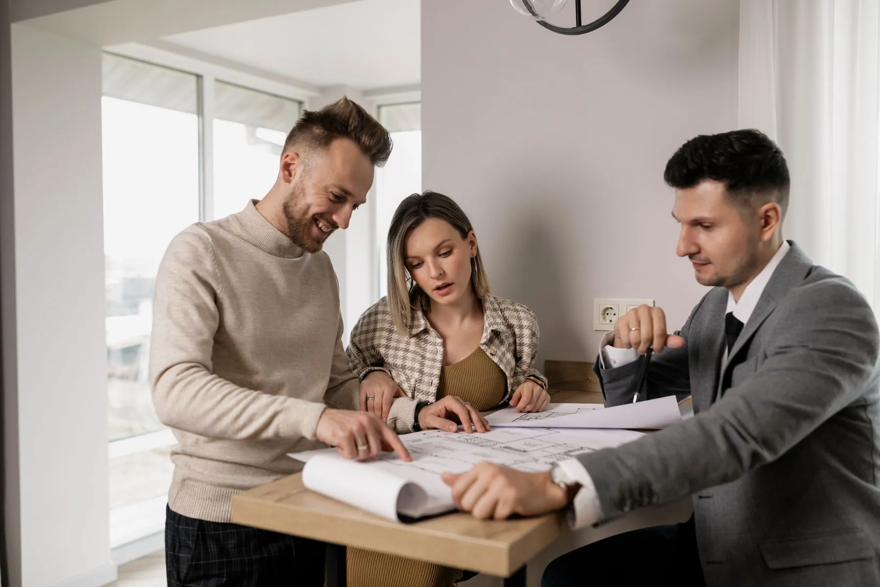 Three people lean over unrolled floor plans spread across a light wooden table.