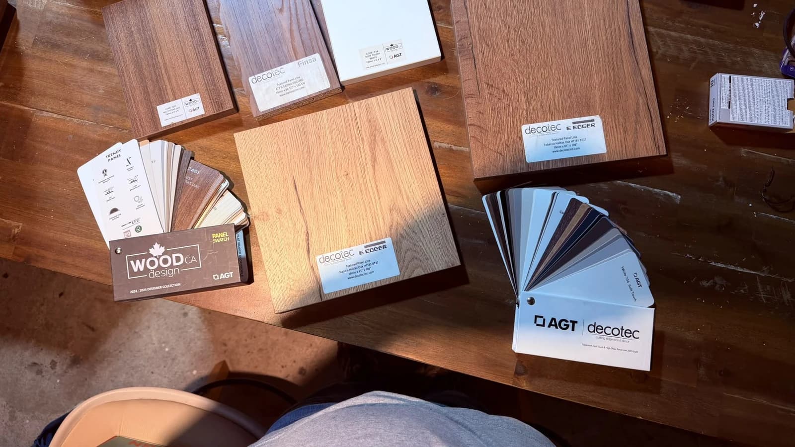 Panel samples laid out on a bench: Egger Halifax Oak boards, a Finsa textured panel, AGT soft-touch white, and the AGT colour fandeck open beside them
